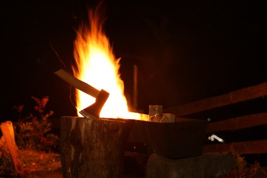 Tree stump with axe and burning firewood in metal brazier outdoors at night