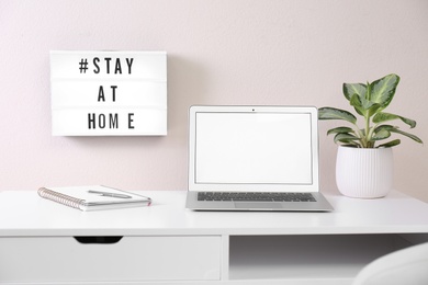 Photo of Laptop, houseplant and lightbox with hashtag STAY AT HOME indoors. Message to promote self-isolation during COVID‑19 pandemic