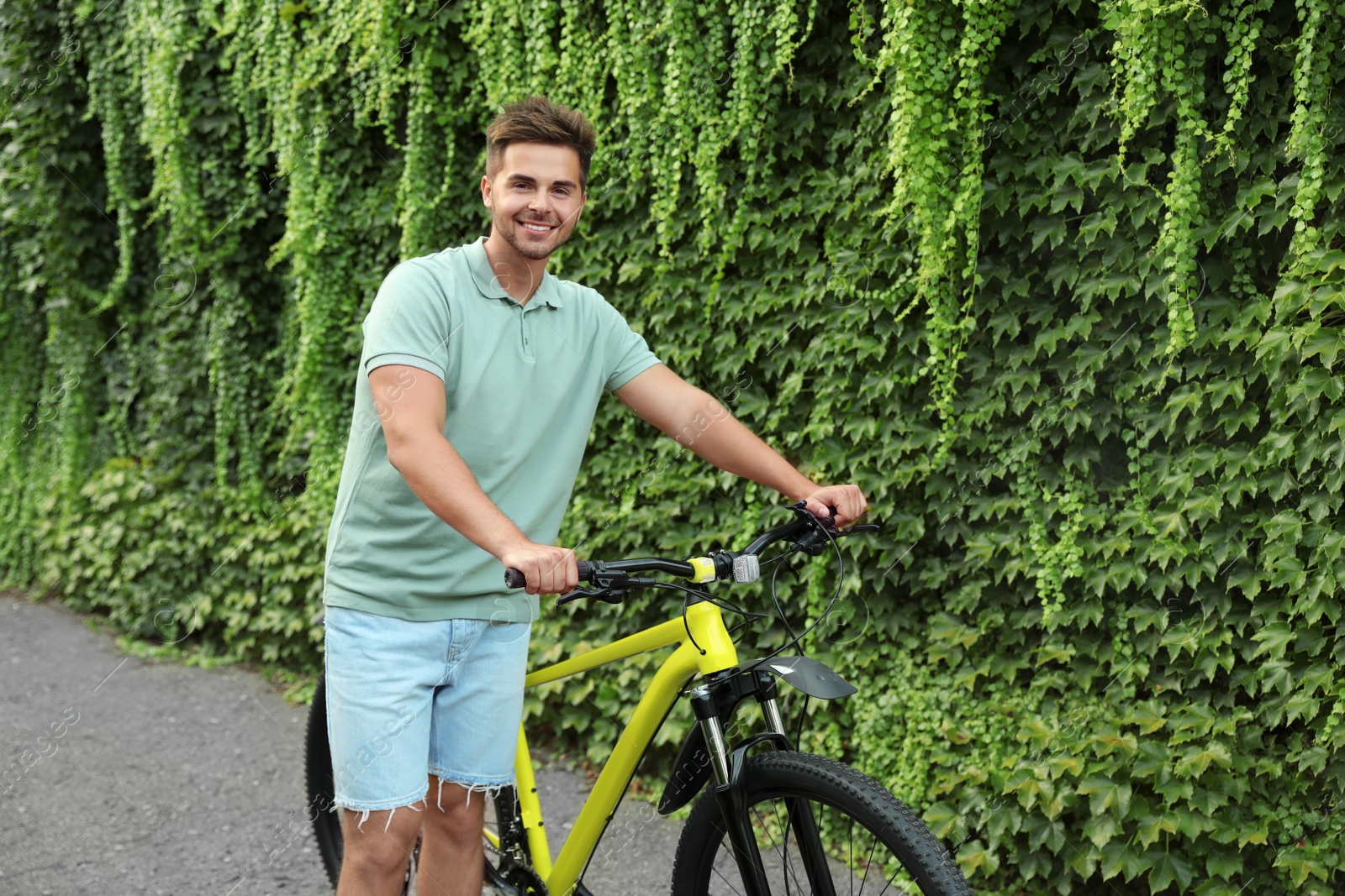 Photo of Handsome young man with bicycle near wall covered with green ivy vines on city street