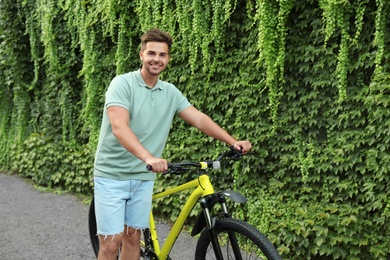 Handsome young man with bicycle near wall covered with green ivy vines on city street