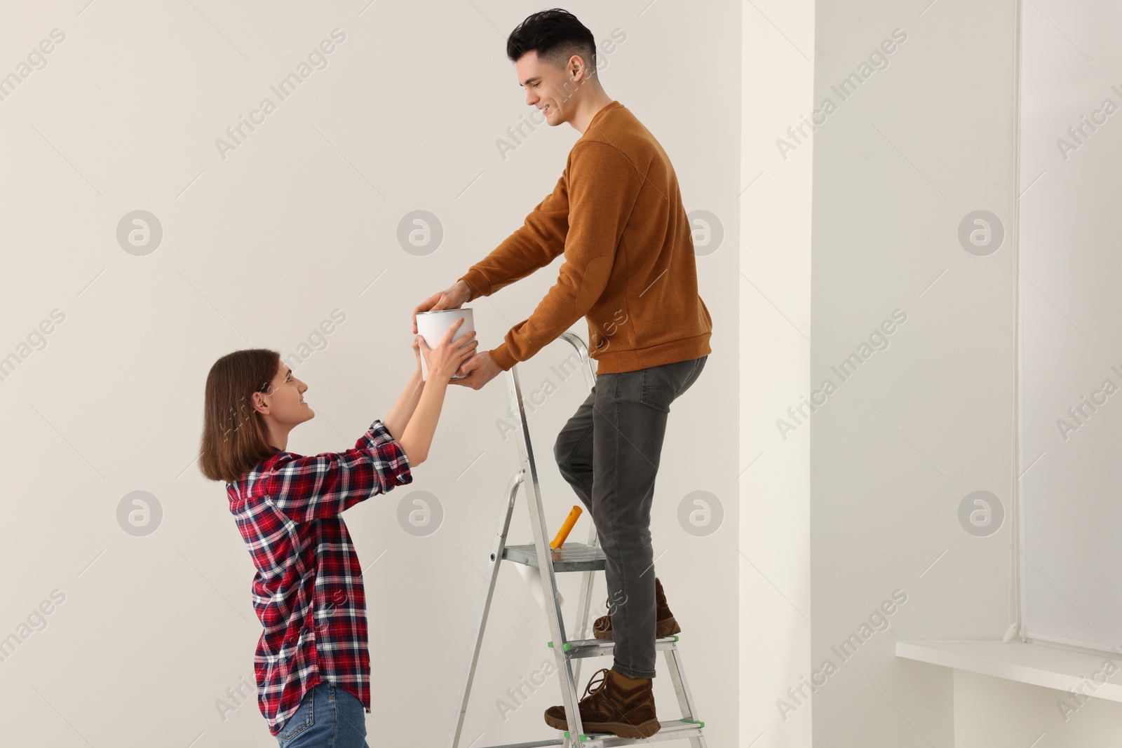 Photo of Young woman giving paint can to boyfriend on stepladder indoors. Room renovation