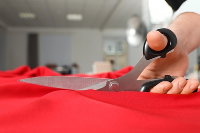 Photo of Professional tailor cutting red fabric with scissors in workshop, closeup