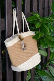 Stylish bag with sunglasses hanging on wooden fence outdoors. Beach accessories