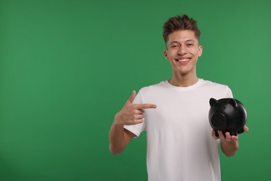 Photo of Happy man pointing at piggy bank on green background. Space for text