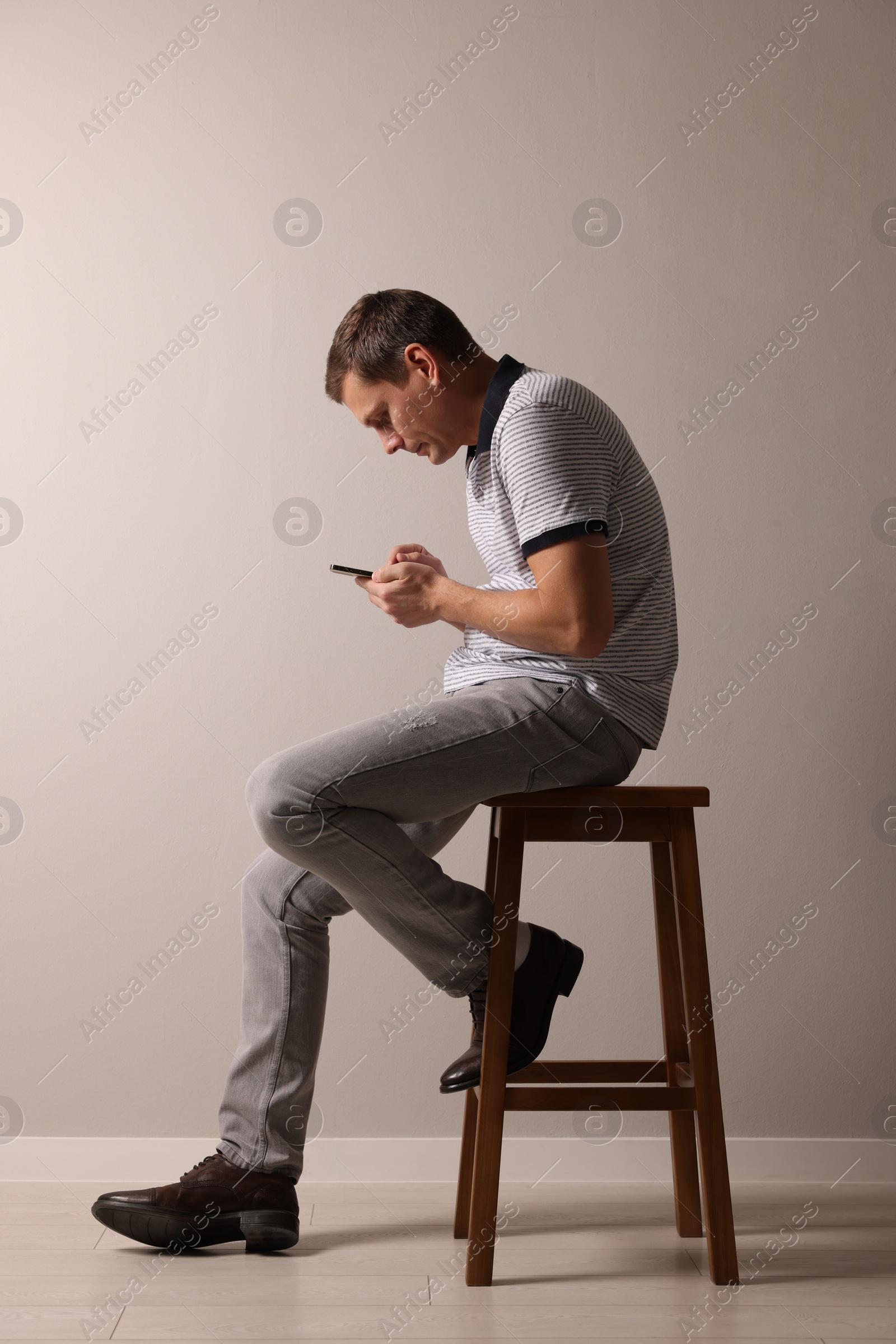 Photo of Man with bad posture using smartphone while sitting on stool against grey background