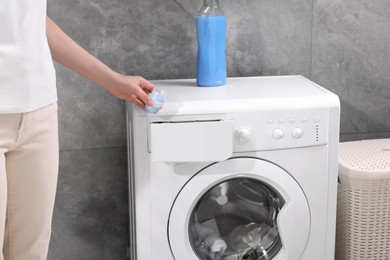 Photo of Woman pouring fabric softener from cap into washing machine near grey wall, closeup