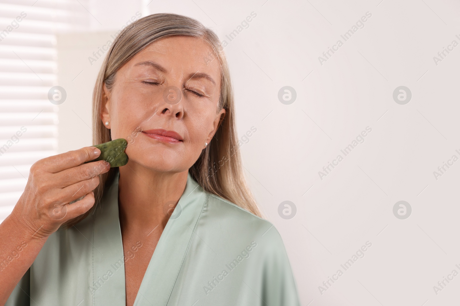 Photo of Woman massaging her face with jade gua sha tool in bathroom. Space for text