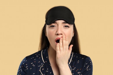 Photo of Tired young woman with sleep mask yawning on beige background. Insomnia problem