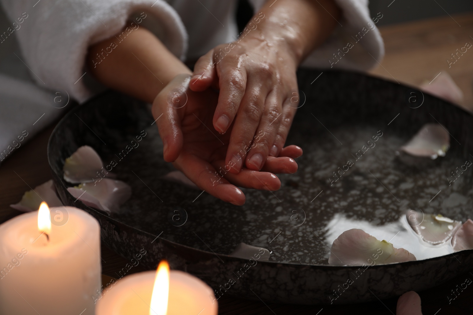 Photo of Woman soaking her hands in bowl of water and flower petals at table, closeup. Spa treatment