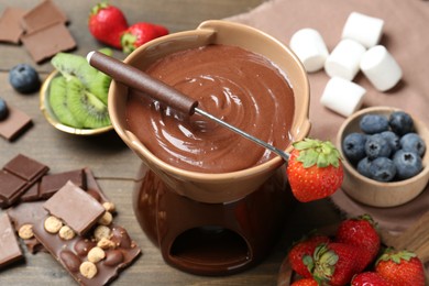 Fondue pot with melted chocolate, different fresh berries, kiwi and fork on wooden table