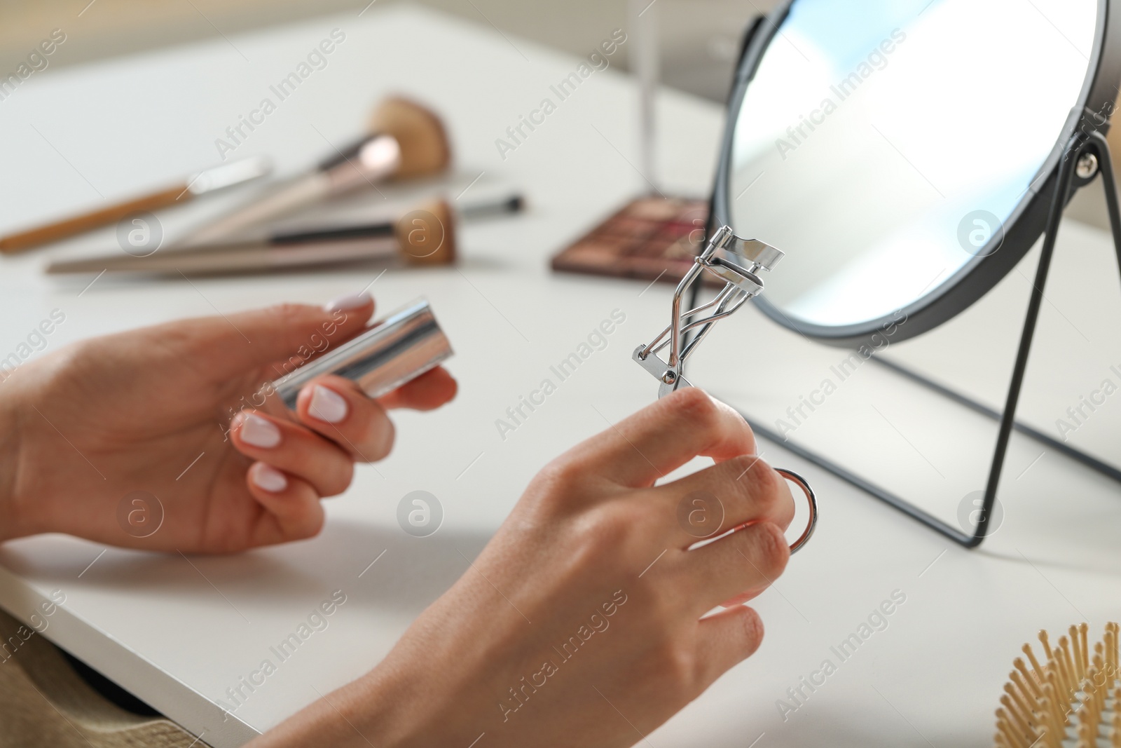 Photo of Woman with eyelash curler, makeup products and mirror at white table, closeup