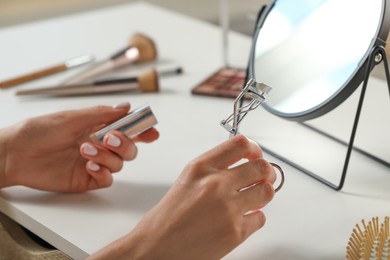 Woman with eyelash curler, makeup products and mirror at white table, closeup