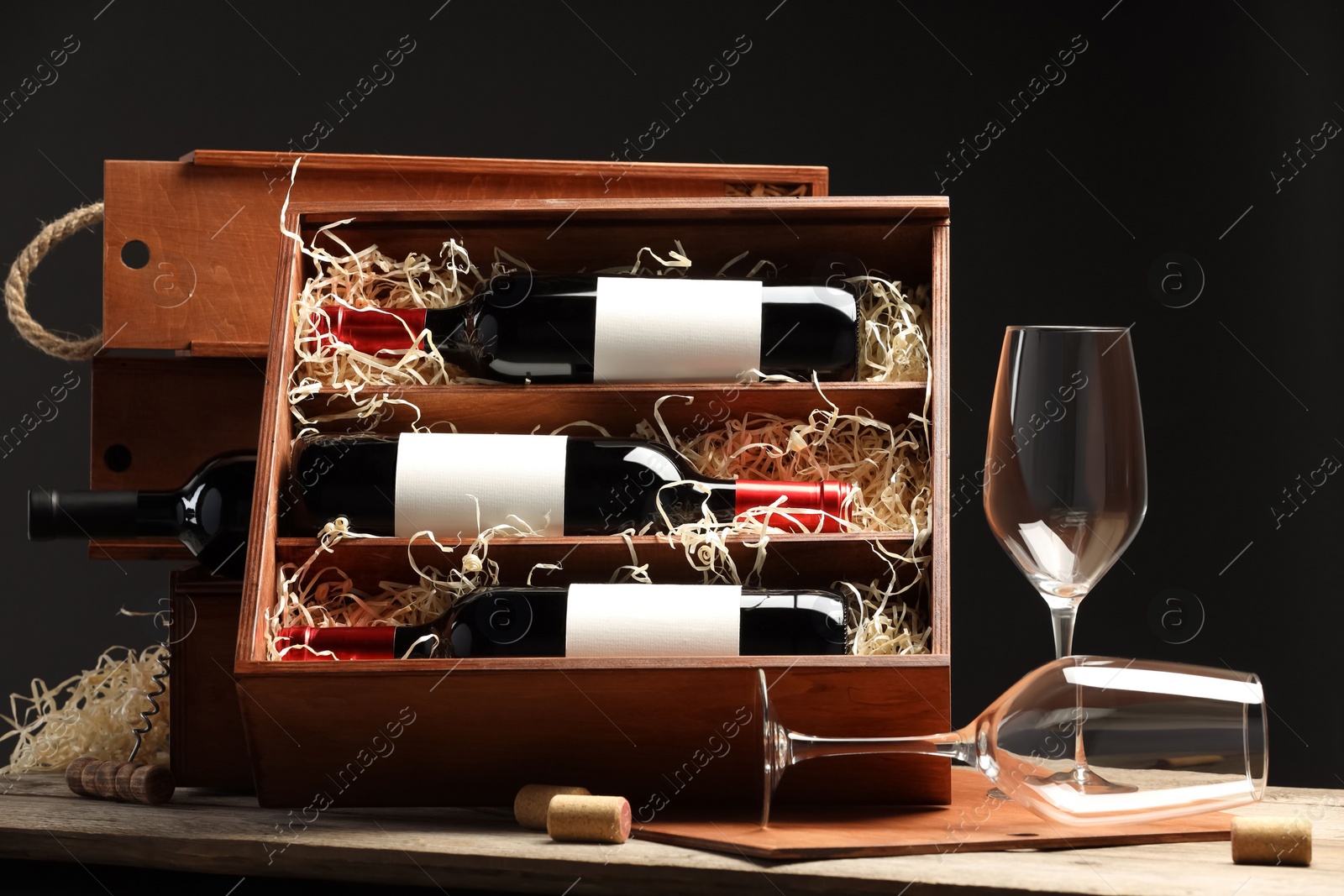 Photo of Box with wine bottles, corks and glasses on wooden table against black background