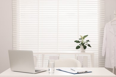 Photo of Doctor's workplace. Laptop, clipboard and glass of water on white table in hospital