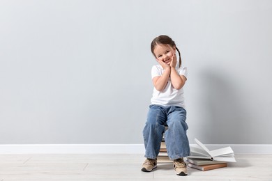 Cute little girl sitting on stack of books near light grey wall. Space for text