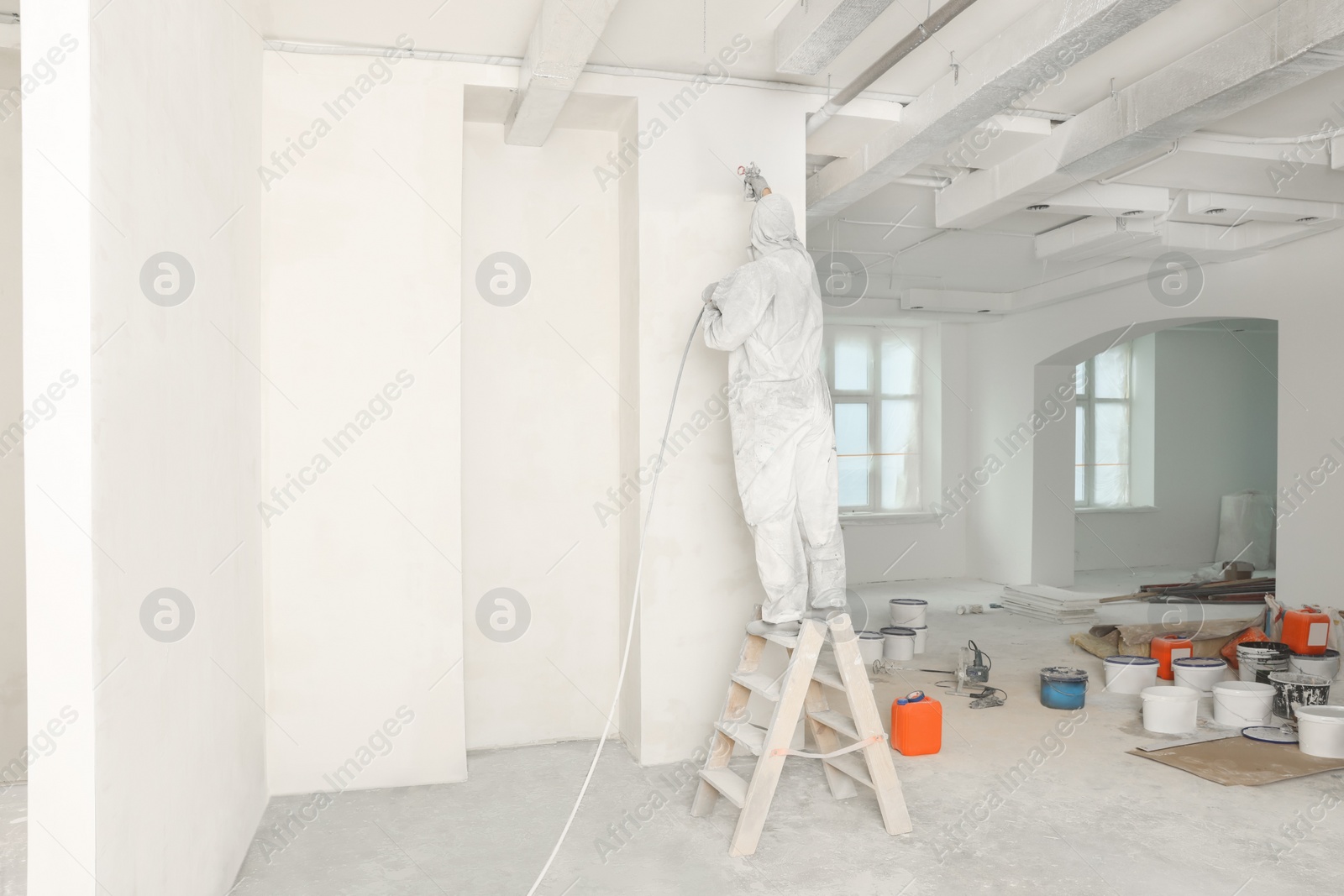 Photo of Decorator in uniform painting wall with sprayer indoors, back view