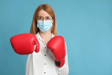 Photo of Woman with protective mask and boxing gloves on light blue background, space for text. Strong immunity concept