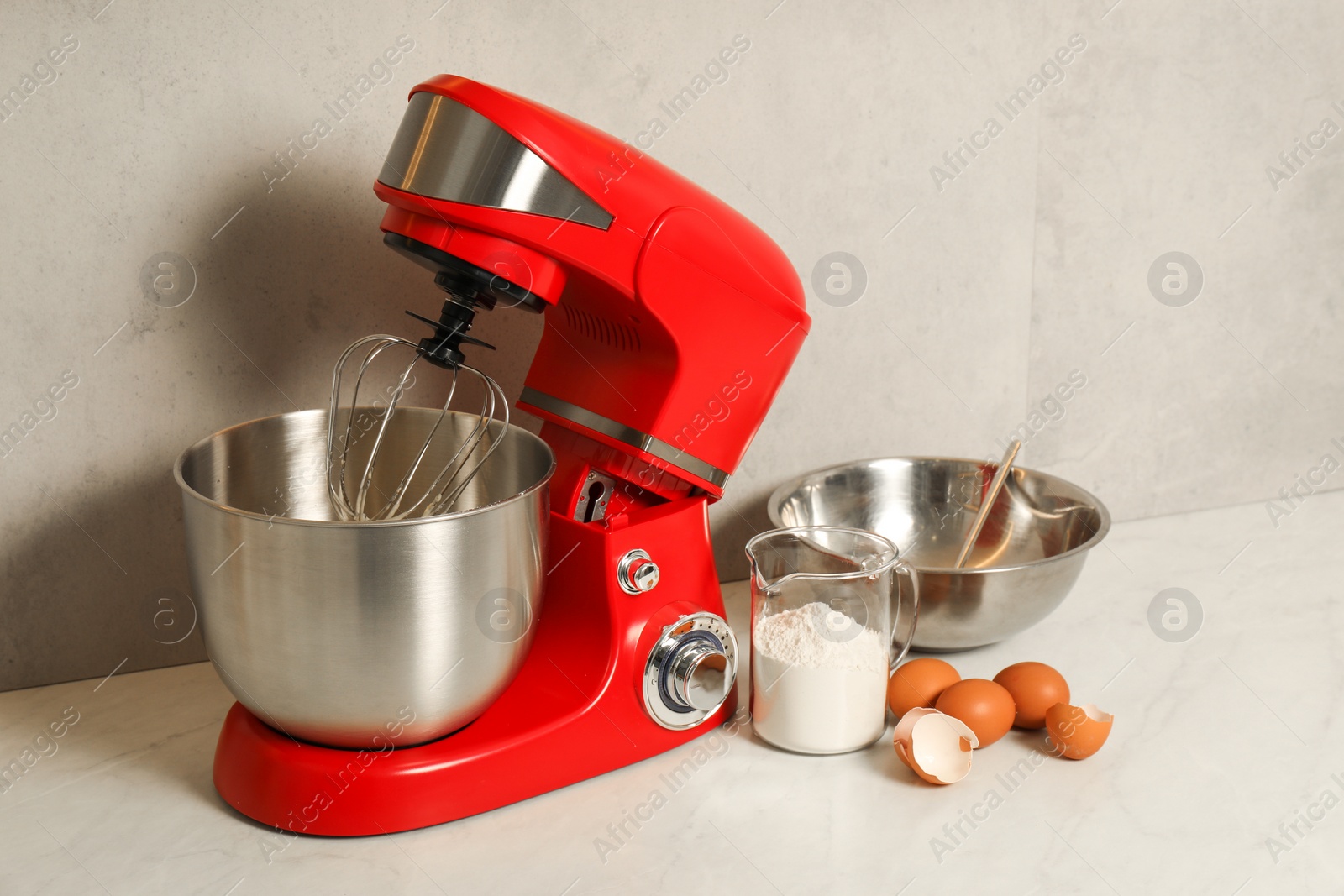 Photo of Modern red stand mixer, eggs, container with flour and bowl on white marble table