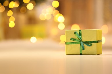 Photo of Beautifully wrapped gift box on white table against blurred festive lights, space for text. Christmas celebration