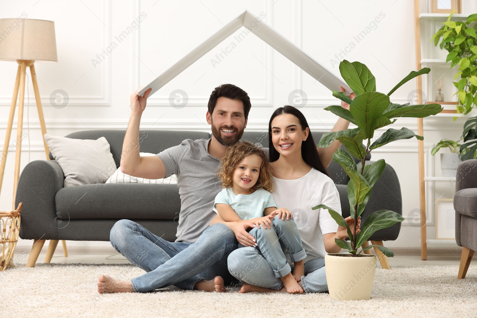 Photo of Housing concept. Happy husband holding plastic roof over his family while sitting on floor at home