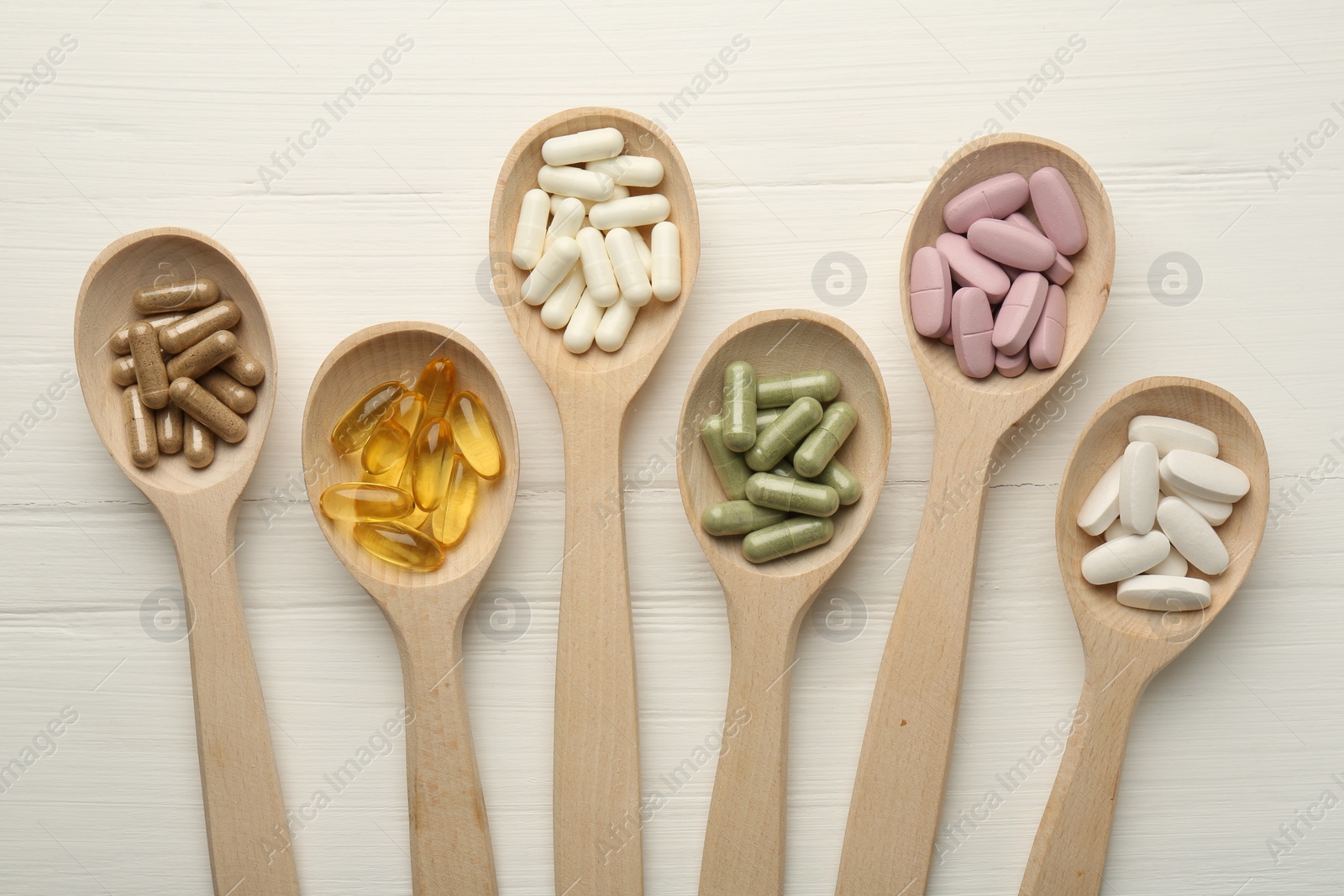 Photo of Different vitamin capsules in spoons on white wooden table, flat lay