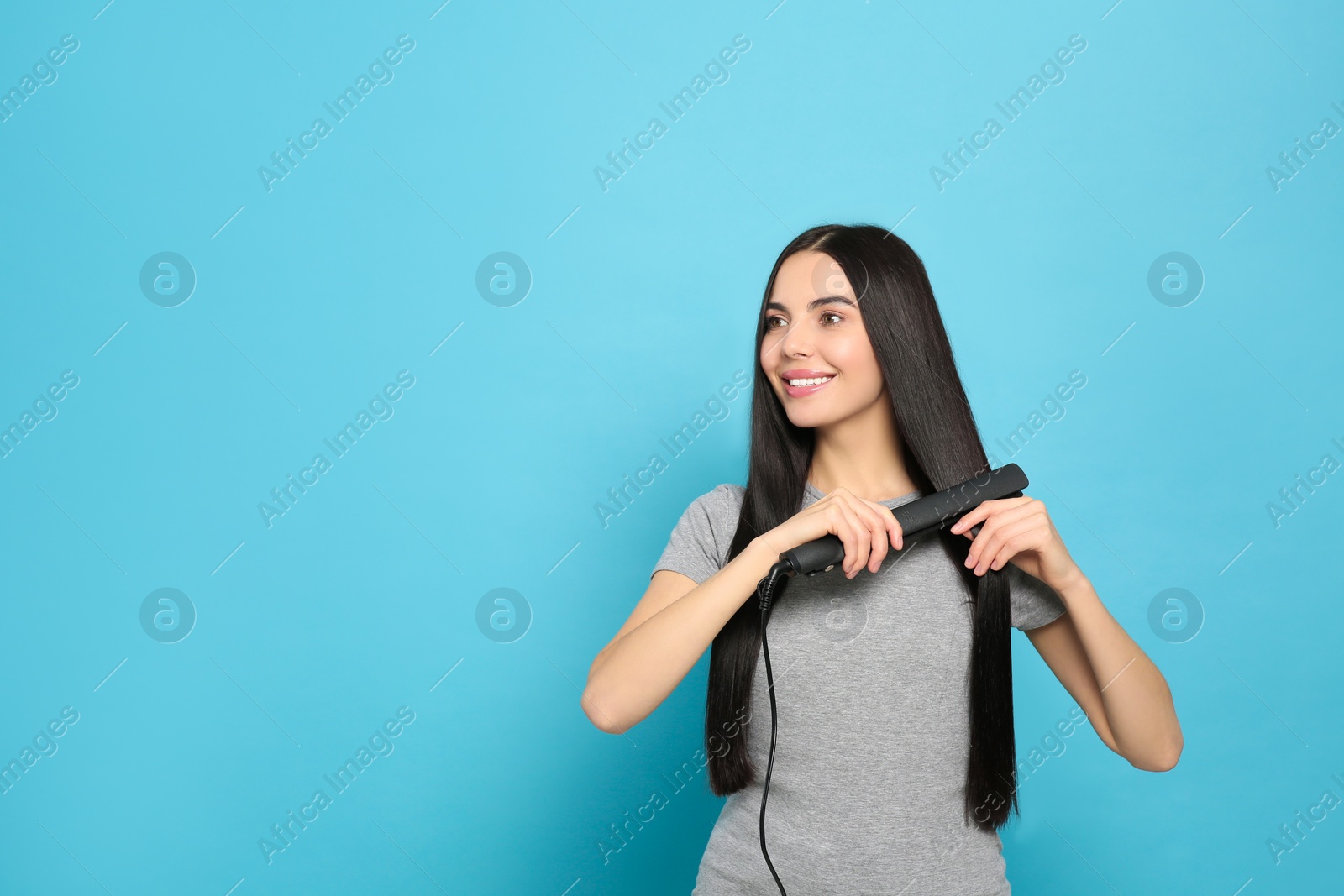 Photo of Beautiful happy woman using hair iron on light blue background. Space for text