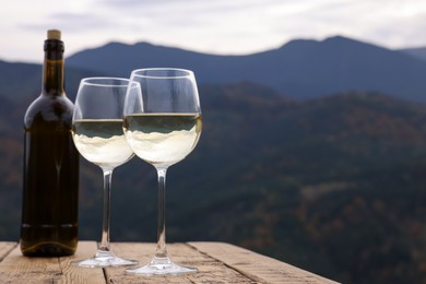 Glasses and bottle of tasty wine on wooden table against mountain landscape. Space for text