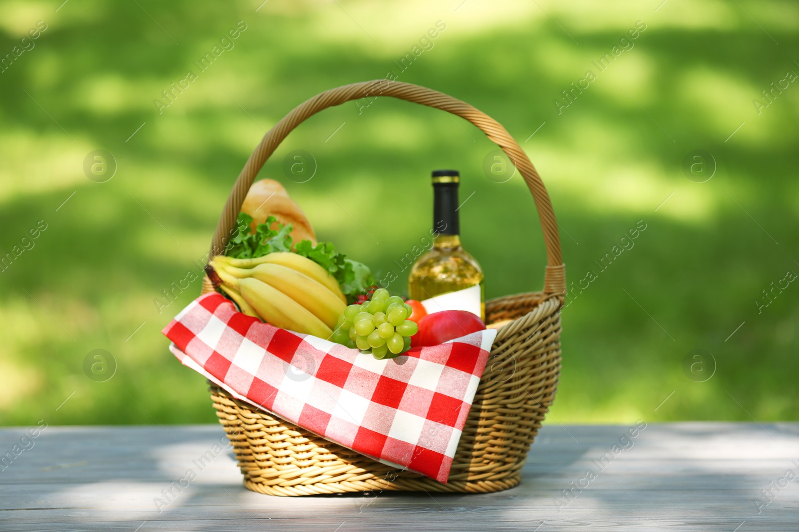 Photo of Wicker basket with blanket, wine and food on table in park. Summer picnic