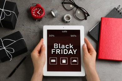 Photo of Woman with tablet surrounded by gifts and accessories at grey table, top view. Black Friday sale