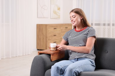 Happy woman holding cup of coffee on sofa armrest wooden table at home, space for text