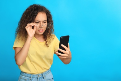 Young African-American woman with vision problems using smartphone on blue background