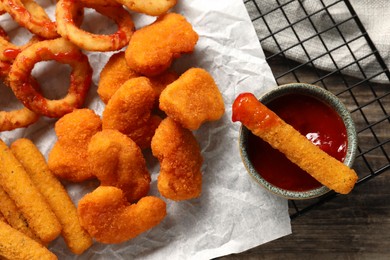 Photo of Tasty chicken nuggets, fried onion rings, cheese sticks and ketchup on wooden table, top view