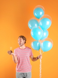 Photo of Young man with birthday muffin and air balloons on color background