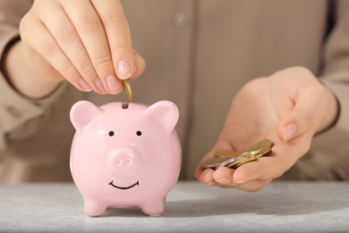 Photo of Pension savings. Woman putting coins into piggy bank at light grey table, closeup