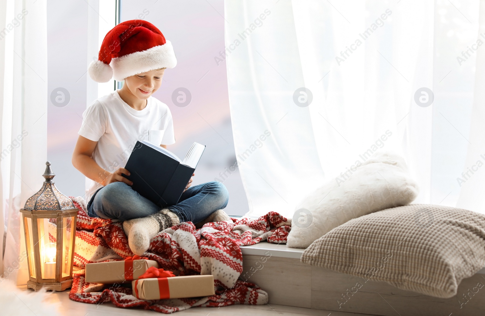 Photo of Cute little child in Santa hat reading Christmas story while sitting on windowsill at home