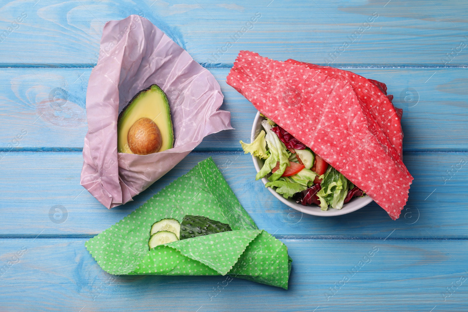 Photo of Different products in beeswax food wraps on light blue wooden table, flat lay