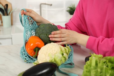 Photo of Woman taking vegetables out from string bag at light marble table, closeup