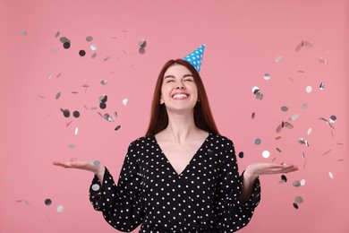 Happy woman in party hat and falling confetti on pink background