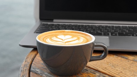 Cup of delicious coffee and laptop on wooden table, closeup