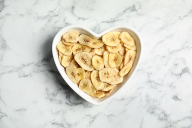 Photo of Heart shaped plate with banana slices on marble table, top view. Dried fruit as healthy snack