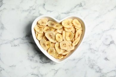 Heart shaped plate with banana slices on marble table, top view. Dried fruit as healthy snack