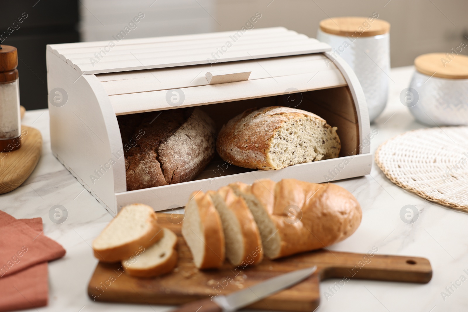 Photo of Wooden bread basket with freshly baked loaves and knife on white marble table in kitchen, closeup