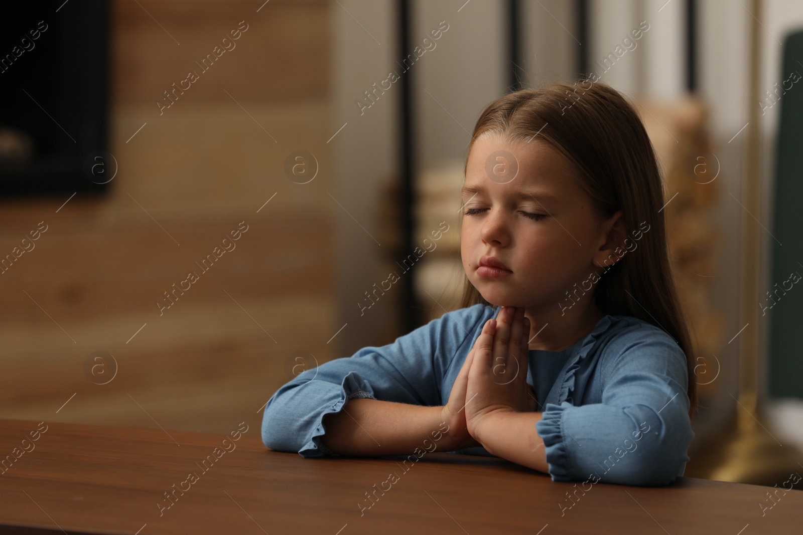 Photo of Cute little girl with hands clasped together praying at table indoors