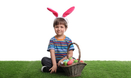 Cute little boy wearing bunny ears with basket full of dyed Easter eggs on green grass