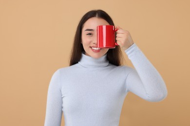Photo of Happy young woman covering eye with red ceramic mug on beige background