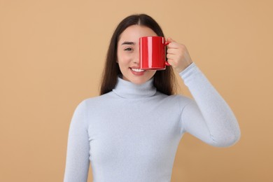 Happy young woman covering eye with red ceramic mug on beige background