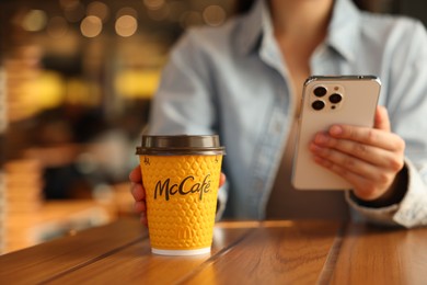 Photo of Lviv, Ukraine - September 26, 2023: Woman with hot McDonald's drink and smartphone at wooden table in cafe, closeup