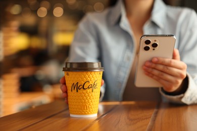 Lviv, Ukraine - September 26, 2023: Woman with hot McDonald's drink and smartphone at wooden table in cafe, closeup