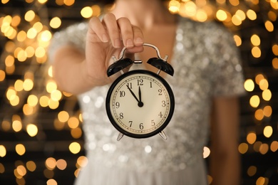 Woman holding alarm clock against blurred lights, closeup. New Year countdown