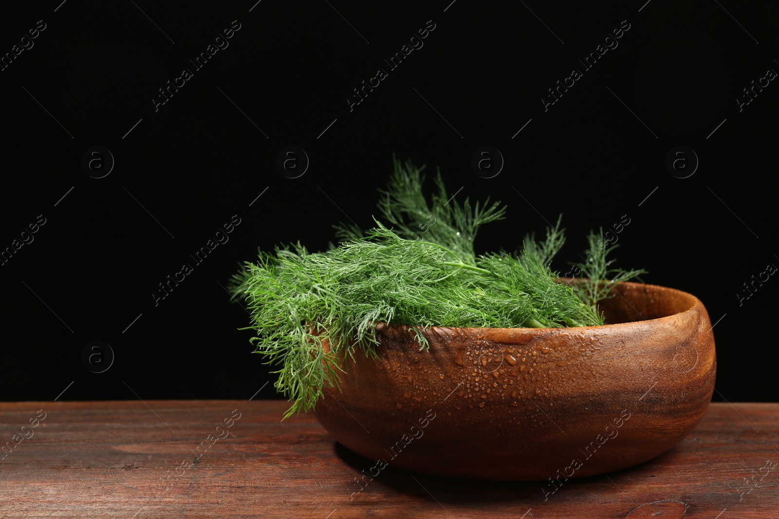 Photo of Bowl of fresh green dill with water drops on wooden table against black background. Space for text